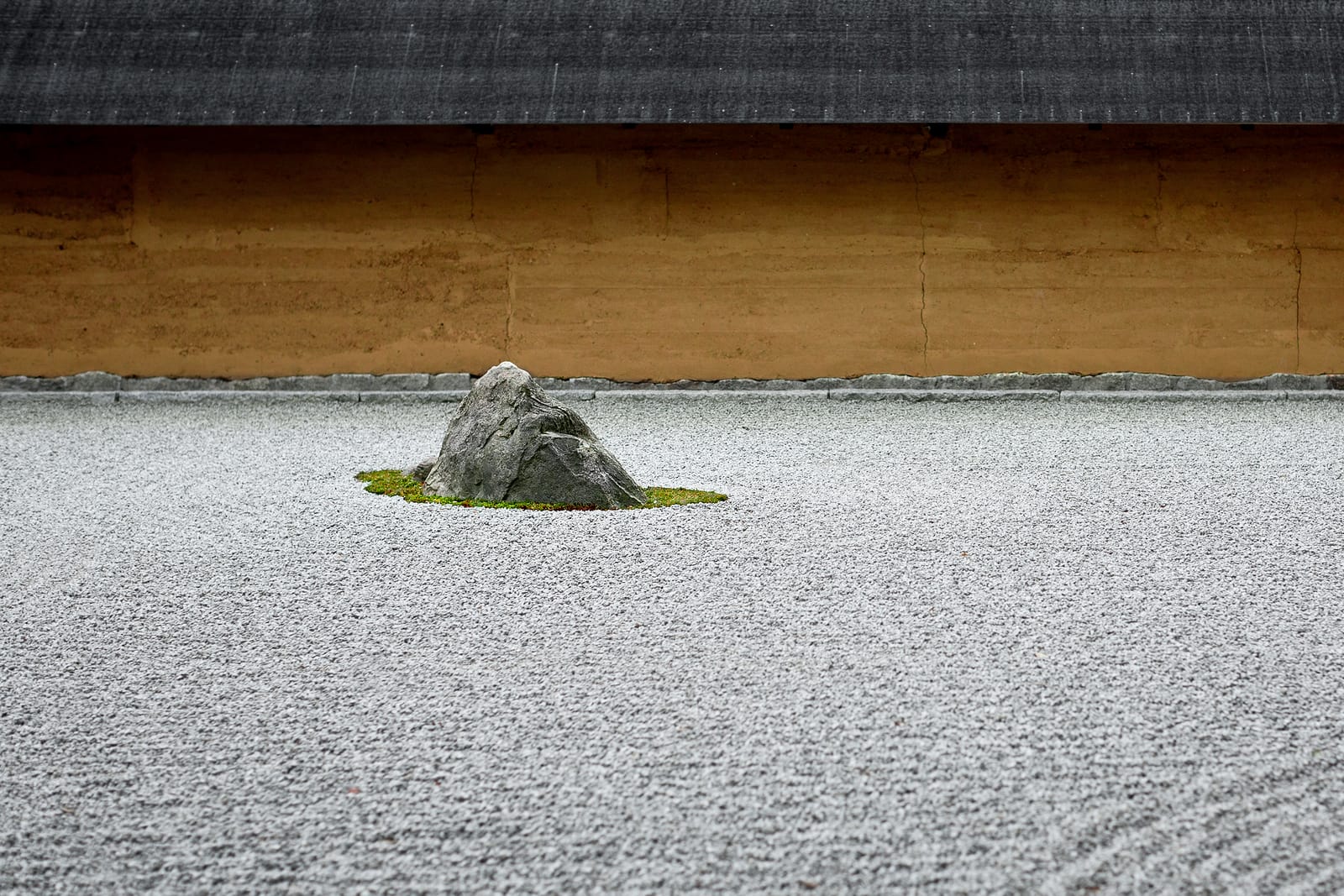 Raked white gravel and a single stone in a Kyoto dry rock garden, an earthen wall and tiled eave behind