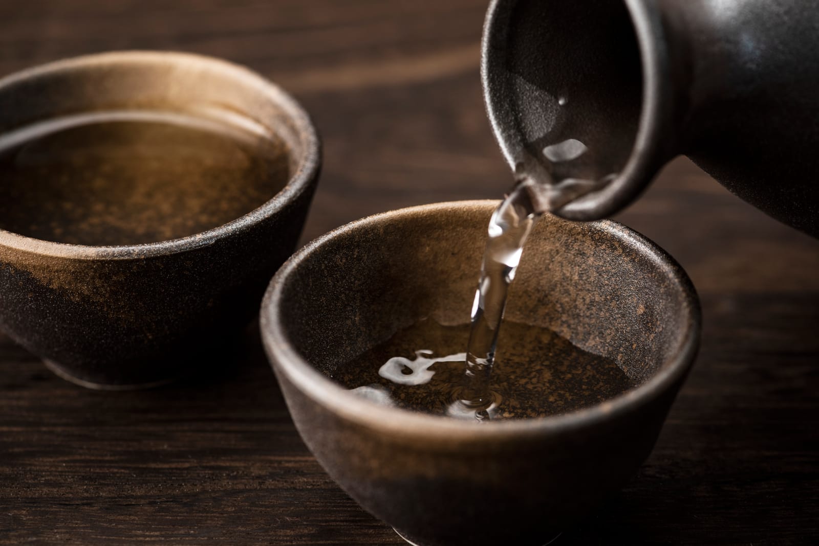 Clear sake being poured from a dark stoneware flask into a matching cup on a wooden table