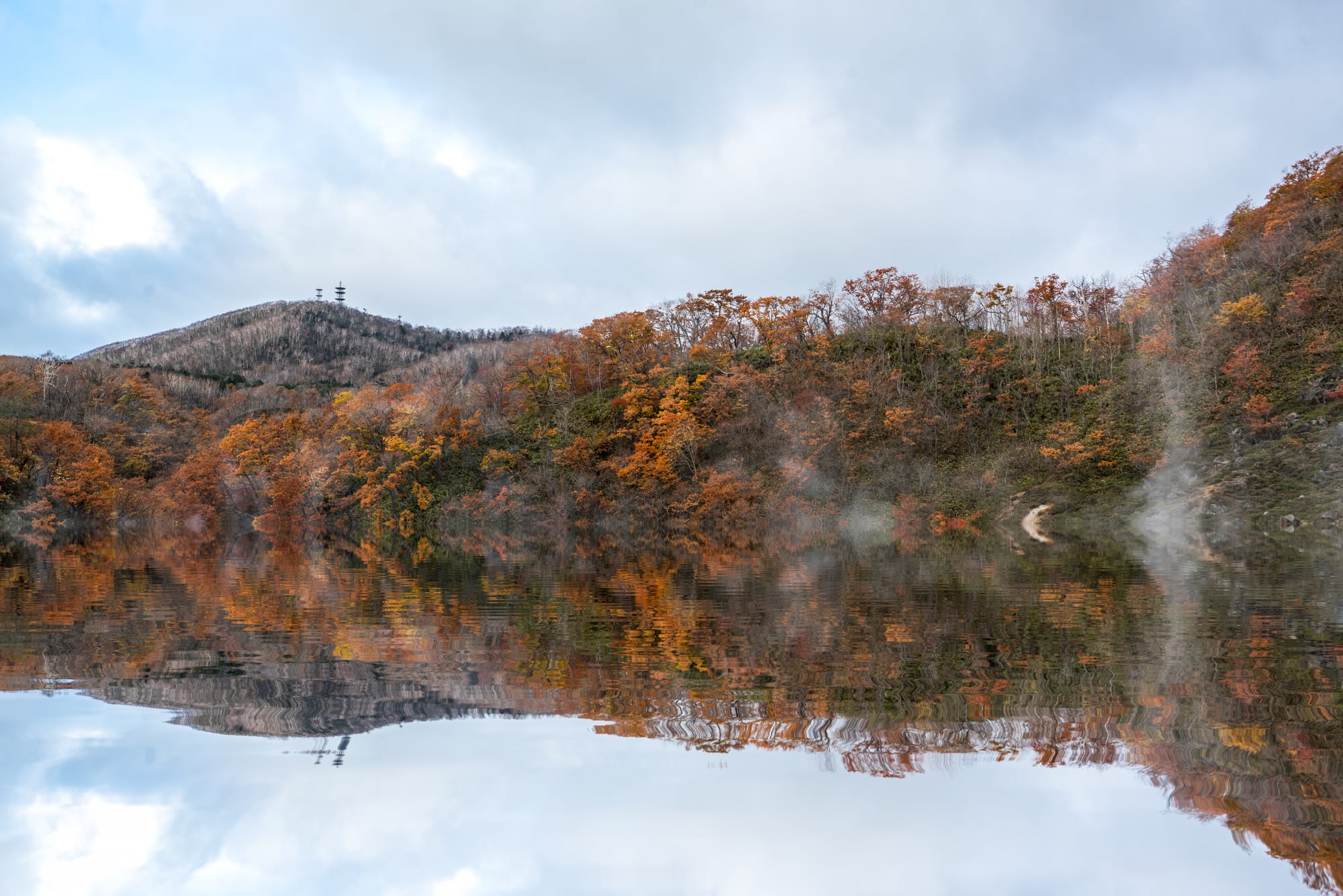 Orange and copper autumn hillsides reflected in a still steaming lake with a distant ridge