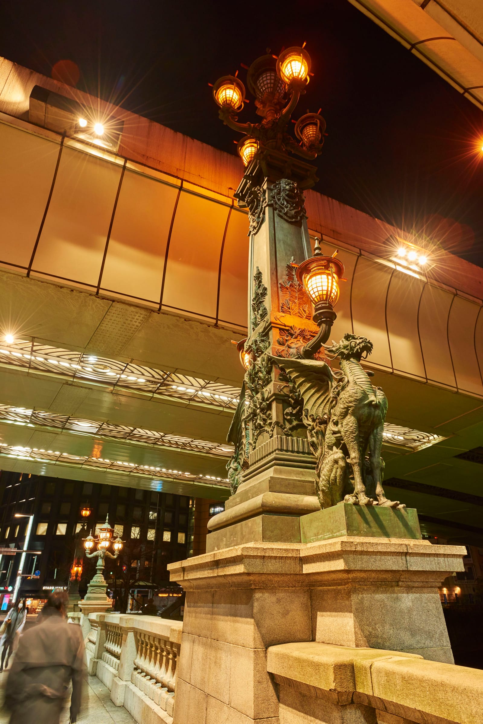 An ornate bronze lamp post on Nihonbashi bridge, its base guarded by a sculpted mythical winged lion-dragon figure, lit at night